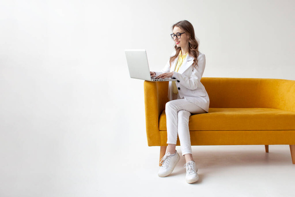 νεαρή επιχειρηματίας με κοστούμι κάθεται σε άνετο μαλακό καναπέ και χρησιμοποιεί φορητό υπολογιστή, το κορίτσι με επίσημη φθορά πληκτρολογεί στον υπολογιστή σε κίτρινο καναπέ σε λευκό απομονωμένο φόντο - Φωτογραφία, εικόνα