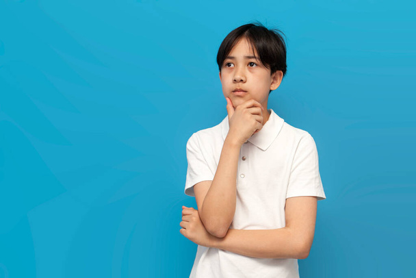 zamyślony azjatycki chłopiec w wieku dwunastu lat myśli i wspomina na niebieskim odosobnionym tle, koreańskie marzenia i plany dzieci i patrzy na przestrzeń kopii - Zdjęcie, obraz