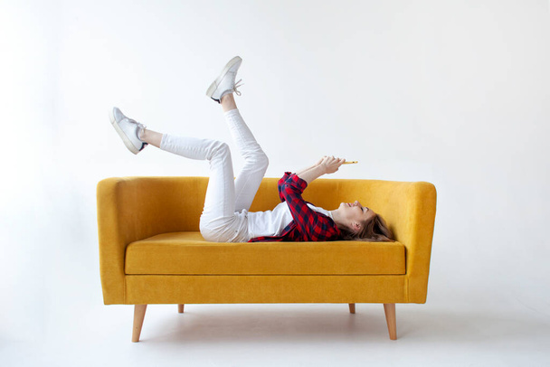 young cute girl lies on soft comfortable sofa with her feet up and uses smartphone, woman communicates by mobile online on yellow couch on white isolated background - Photo, Image