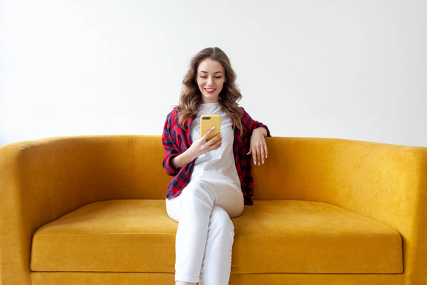 junges nettes Mädchen nutzt Smartphone auf weichem bequemen Sofa, Frau tippt Nachricht auf Handy online auf gelbem Sofa auf weißem isolierten Hintergrund - Foto, Bild