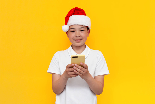little asian boy in santa hat uses smartphone on yellow isolated background, korean child in new year's hat types online on the phone for christmas - Photo, image