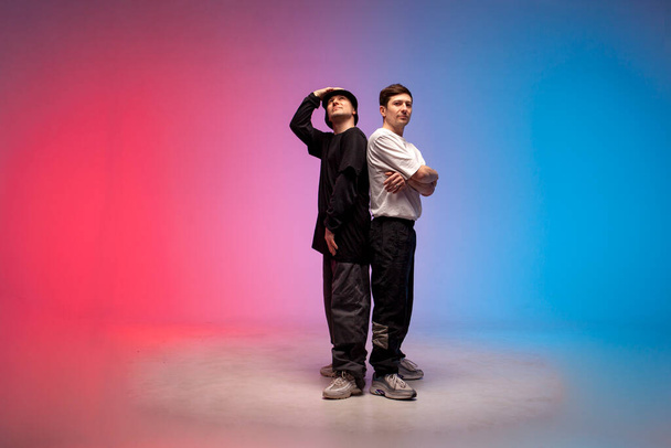 two dancer guys stand together in neon club lighting, male rap performers stand on red blue background and look up - Photo, Image