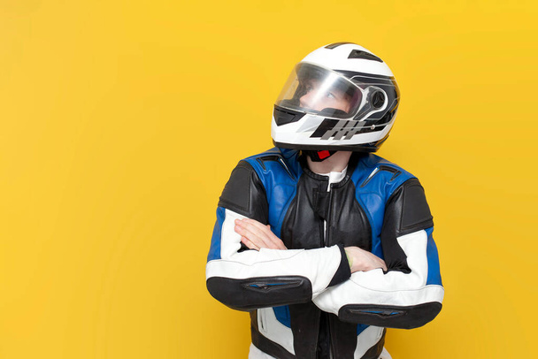 man motorrijder in lederen jas en helm staat met zijn armen gekruist op geel geïsoleerde achtergrond en kijkt weg naar de ruimte om te gaan, motorfiets racer in beschermende versnelling adverteert lege ruimte - Foto, afbeelding