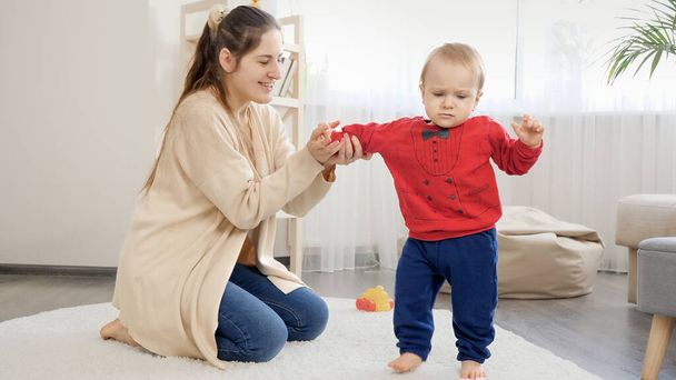 Smiling young woman support and holding her baby son learning walking on carpet at living room. Baby development, family playing games, making first steps, parenthood and care. - Photo, Image