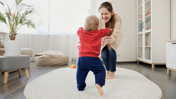 1歳の男の子は歩くことを学び、母親を笑わせるためにカーペットの上でステップを作ります。赤ちゃんの発達、家族で遊ぶゲーム、第一歩を踏み出す、親とケア. - 写真・画像