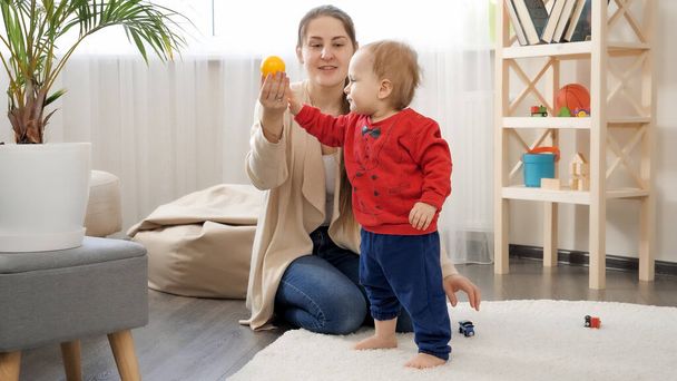 Pikkupoika ottaa värikkäitä leluja äidiltään ja kävelee pehmeällä matolla olohuoneessa. Vauvan kehitys, perheen pelit, ensiaskeleet, vanhemmuus ja hoito. - Valokuva, kuva