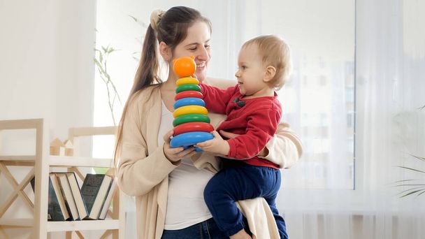 Sonriente madre sosteniendo su bebé som y dándole colorida torre de juguete o piramida. - Foto, imagen