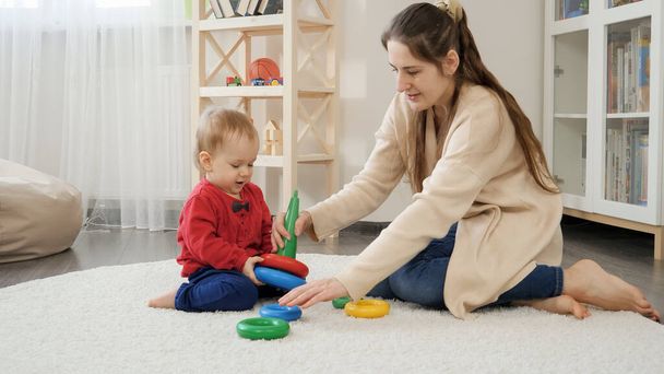 Beautiful young mother playing with little baby son on floor and assembling toy tower. Baby development, child playing games, education and learning - Foto, immagini