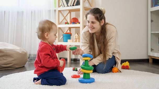Sorridente madre giocando con il suo bambino figlio sul tappeto e dandogli giocattoli colorati. - Foto, immagini