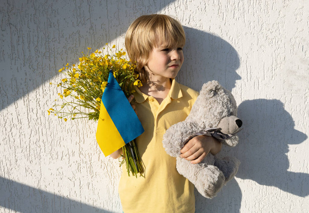dramatic portrait of Ukrainian boy holding teddy bear and yellow-blue flag . contrast of light and shadows on wall. War and social problems in Ukraine. loneliness, sadness and nostalgia for Ukraine - Photo, image