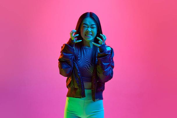 Anger, irritation. Portrait of young korean girl in casual clothes posing against pink studio background in neon light. Concept of emotions, facial expression, youth, lifestyle, news, ad - 写真・画像
