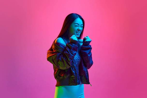 怒り、刺激。ネオンの光の中でピンクのスタジオを背景に拳でポーズを取るカジュアルな服の中に若い韓国人の少女の肖像画。感情の概念,顔の表情,若者,ライフスタイル,広告 - 写真・画像
