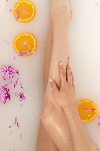 Portakal dilimleri ve çiçek yapraklarıyla dolu bir banyoda narin kız bacakları. Güzellik ve cilt bakımı. Üst görünüm. - Fotoğraf, Görsel