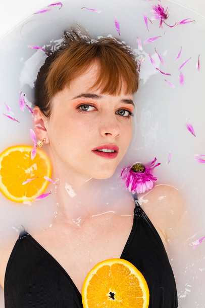 Gyengéd lány fekszik a fürdőben félig vízben, tele gyümölcsös narancsszeletekkel és virágszirmokkal. Szépség és bőrápolás koncepció. Felülnézet. - Fotó, kép