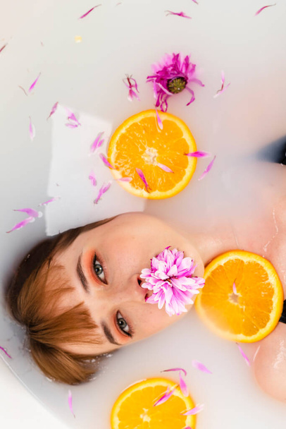 Arka tyttö makaa kylvyssä puoli vedessä täynnä hedelmiä appelsiini viipaleita ja kukka terälehtiä. Kauneus ja ihonhoito käsite. Huippunäkymä. Pää ammuttu. - Valokuva, kuva