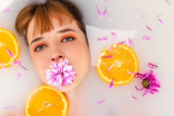 Τρυφερή κοπέλα ξαπλωμένη στο μπάνιο μισογεμάτη νερό με φέτες πορτοκαλιού φρούτων και πέταλα λουλουδιών. Ομορφιά και φροντίδα του δέρματος έννοια. Στο πάνω μέρος. Πυροβολισμός στο κεφάλι. - Φωτογραφία, εικόνα