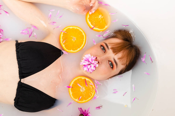 Zacht meisje ligt in een bad half in water gevuld met fruit sinaasappelschijfjes en bloemblaadjes. Schoonheid en huidverzorging concept. Bovenaanzicht. hoofdschot. - Foto, afbeelding