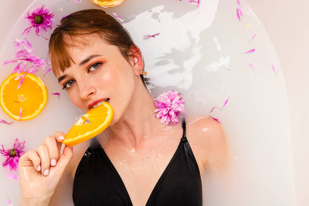 Zacht meisje ligt in een bad half in water gevuld met fruit sinaasappelschijfjes en bloemblaadjes. Schoonheid en huidverzorging concept. Bovenaanzicht. hoofdschot. - Foto, afbeelding