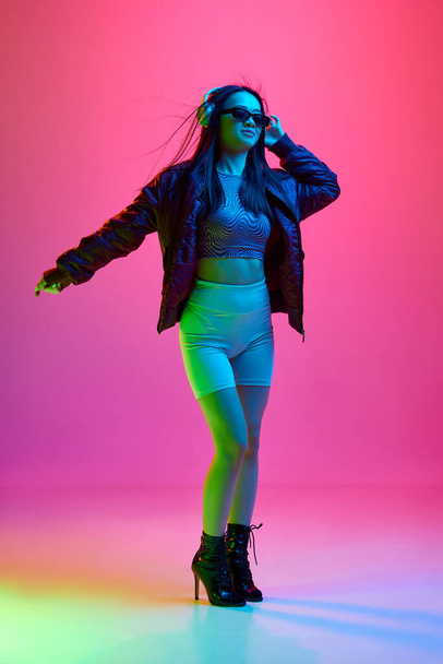 Portrait of pretty, young girl in sunglasses, listening to music in headphones against pink studio background in neon light. Concept of emotions, facial expression, youth, lifestyle, inspiration, ad - Photo, Image