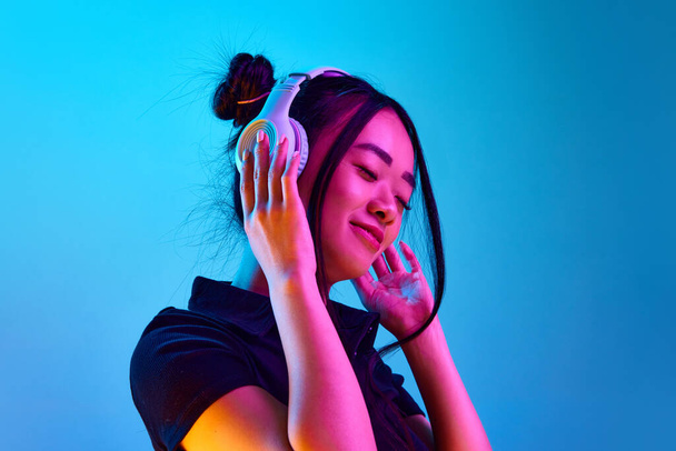 Portré gyönyörű fiatal koreai lány hallgat zenét fülhallgató ellen kék stúdió háttér neon fényben. Fogalom érzelmek, arckifejezés, fiatalok, életmód, inspiráció, értékesítés, hirdetés - Fotó, kép