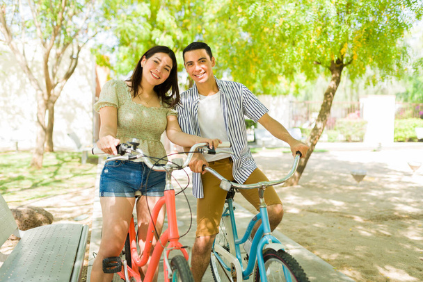Bella coppia di trentenni che guarda la fotocamera sorridente mentre guida le loro biciclette vintage nel parco durante un appuntamento  - Foto, immagini
