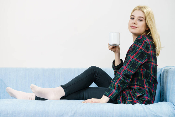 Pretty hipster young adult teenage woman sitting on sofa enjoying her leisure time drinking coffee or tea from mug. - Foto, immagini