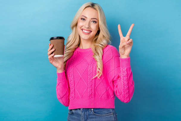 Portrait of satisfied gorgeous woman with curly hairstyle wear knit pullover hold drink showing v-sign isolated on blue color background. - Photo, Image
