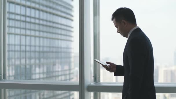 ázsiai üzletember nézi mobiltelefon miközben járkál az ablak az irodában - Felvétel, videó