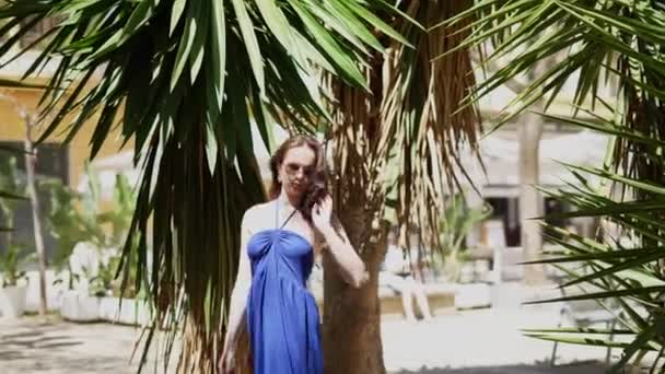 Una ragazza in abito blu cammina lungo un vicolo di palme in una luminosa giornata di sole. Filmati 4k di alta qualità - Filmati, video