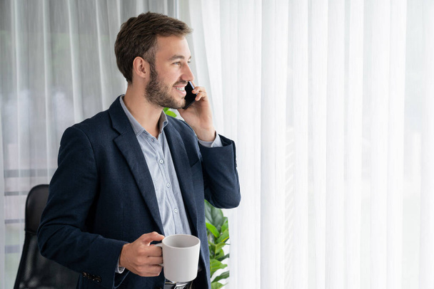 Ciężko pracujący biznesmen stoi pewnie w nowoczesnym biurze, wykonując przekonywujący telefon do klienta. Pracownik biurowy rozmawia przez telefon koordynować i zarządzać pracą biznesową z kolegami. Podmiot - Zdjęcie, obraz