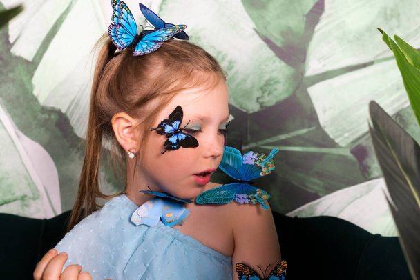 Schattig klein meisje in blauwe jurk met vlinders op haar tussen de planten en grote bladeren. - Foto, afbeelding