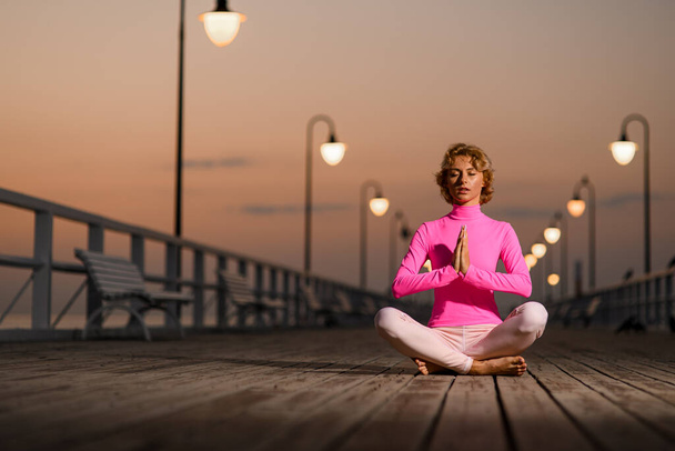 Front view of young woman doing yoga near seaside in the evening. Girl sitting in lotus position on a wooden quay. - Foto, Imagen