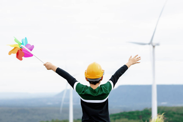 Progressive young asian boy playing with wind pinwheel toy in the wind turbine farm, green field over the hill. Green energy from renewable electric wind generator. Windmill in the countryside concept - Foto, Imagem