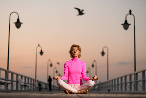 Young blonde woman sitting in lotus pose and slowly breathing during meditation on wooden quay near seashore - Foto, immagini