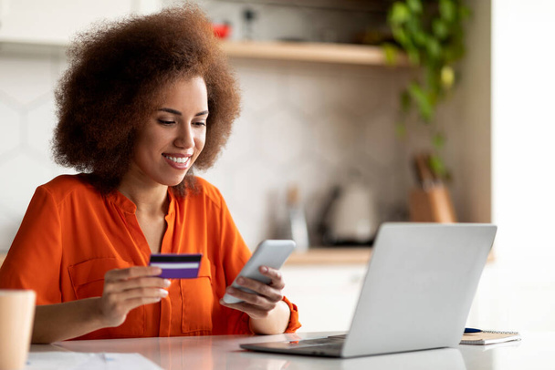 Çevrimiçi ödemeler. Akıllı telefon ve kredi kartı kullanan siyahi bir kadın merkez büroda dizüstü bilgisayarla otururken gülümseyen Afrikalı Amerikalı bir kadın faturaları internet üzerinden ödüyor ya da uygulamayla para transfer ediyor. - Fotoğraf, Görsel