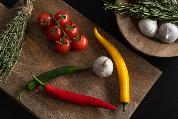 Multicolored pods of fresh hot chili peppers lie on a wooden textured  cutting board on a black  table. Nearby lie garlic, cherry tomato branch, rosemary sprigs. Dark photo, top view.  Healthy lifestyle concept. - Fotografie, Obrázek