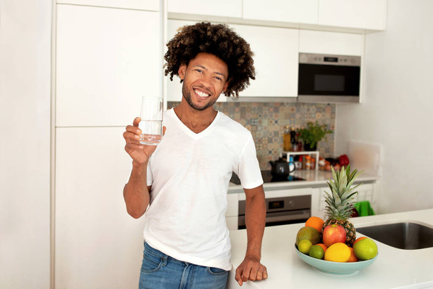Healthy lifestyle. Happy black guy holding glass of water advertising proper hydration for weight loss standing in modern kitchen interior indoor. Stay fresh and hydrated concept - Photo, Image