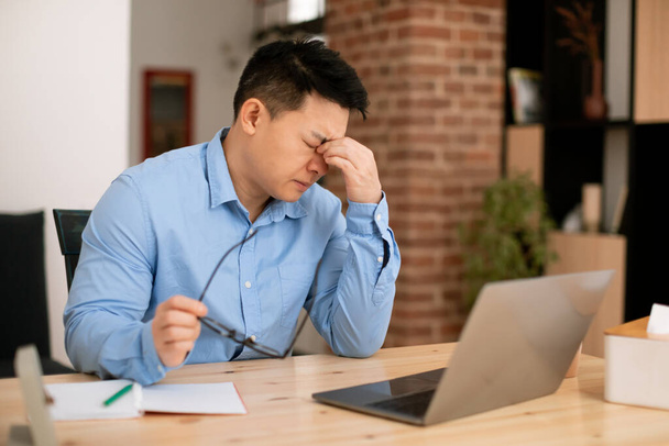 Zmęczony azjatycki w średnim wieku wolny strzelec czuje napięcie oczu po pracy na laptopie zdalnie z domu. Wyczerpany mężczyzna siedzący przy biurku z komputerem i masującym mostem nosowym - Zdjęcie, obraz