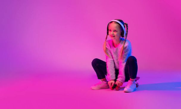 Happy cheerful pretty school aged girl in casual outfit sitting on skateboard on luminous background, looking at copy space and smiling, using wireless headphones, listening to music, web-banner - Фото, зображення
