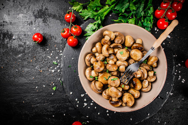 Fried mushrooms in a bowl with greens and tomatoes on a stone board. On a black background. High quality photo - Fotó, kép