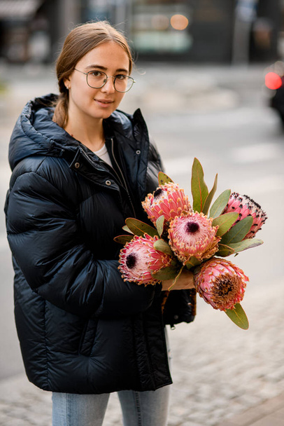 Beautiful girl standing in the street and holding in hands fresh bouquet of pink Protea artichoke flowers. Blurred background - Foto, Imagem