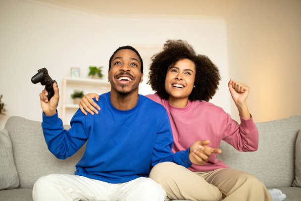 Digitale Unterhaltung. Glückliche junge schwarze Ehepartner feiern den Sieg beim Videospiel, schütteln die Fäuste und halten den Gamepad-Controller auf der Couch zu Hause. Wochenendspaß-Konzept - Foto, Bild