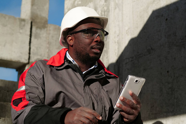 ハード帽子のアフリカ系アメリカ人労働者とスマートフォンで保護ゴーグルは建設現場に座っています。ワークマンは白いスマートフォンを手に持っています - 写真・画像