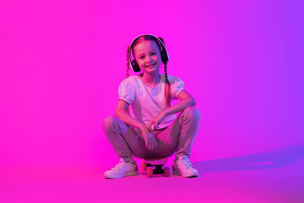 Skateboarding Prodigy, childhood concept. Cheerful pretty little girl skateboarder practicing skills on futuristic background, smiling at camera, using wireless headphones, full length, copy space - Foto, imagen