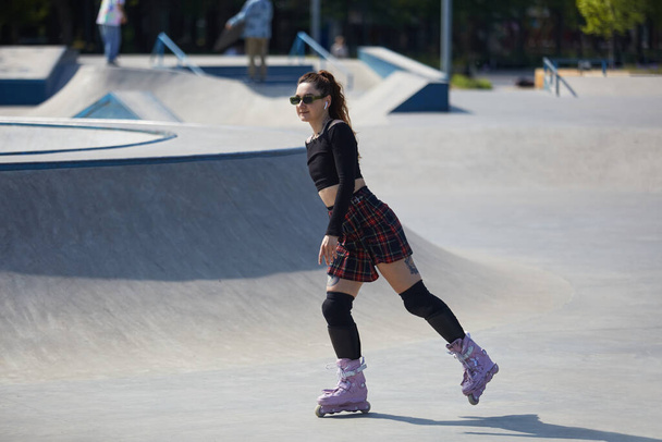 Cute young adult female riding on aggressive inline skates in a concrete skatepark. Roller blader chick in plaid skirt skating in a urban park in summer - Zdjęcie, obraz