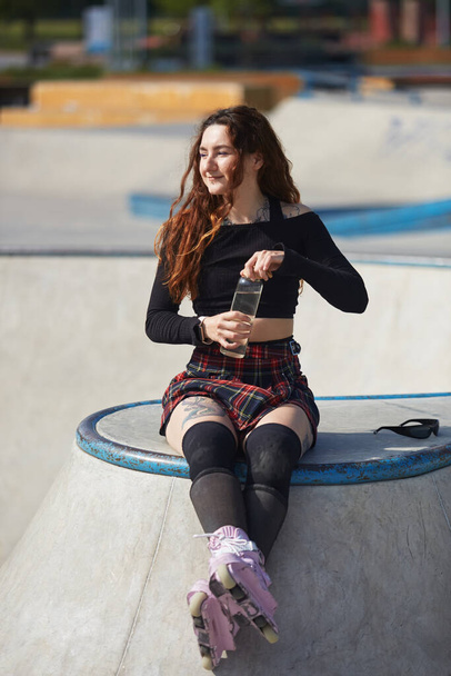 Aggressive inline skater female wearing modern purple roller blades drinking water from a reusable glass bottle. Cool tattooed young person resting in a skatepark in a sunny summer day - Foto, immagini