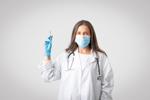 Σοβαρή ηλικιωμένη γιατρός με λευκό παλτό, προστατευτική μάσκα που κρατά σύριγγα με εμβόλιο, στέκεται σε γκρι φόντο στούντιο. Ανοσοποίηση για υγειονομική περίθαλψη, θεραπεία ασθενειών και εμβολιασμό - Φωτογραφία, εικόνα