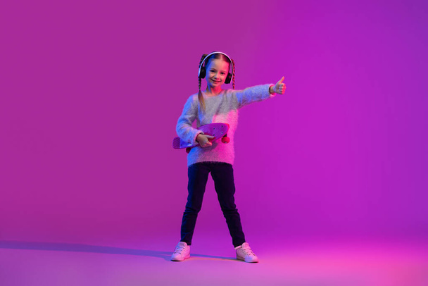 Generation alpha. Happy cheerful cute preteen stylish girl having fun on luminous background, holding skateboard, using wireless headphons, showing thumb up and smiling, copy space - Photo, Image
