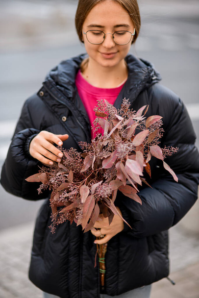 Young attractive girl florist standing on the street and holding bouquet with dark red eucalyptus Populus branches in hands on blurred background - Photo, Image