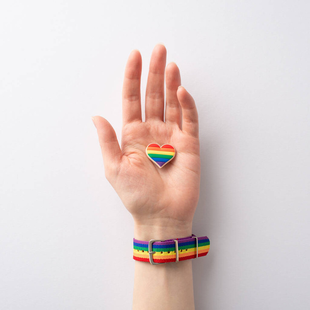 Een eerste persoon bovenste vierkante weergave van symbolische regenboog gekleurde armband siert een vrouwelijke hand met een hartvormige pin badge op een witte achtergrond, ter ere van LGBTQ Geschiedenis Maand - Foto, afbeelding